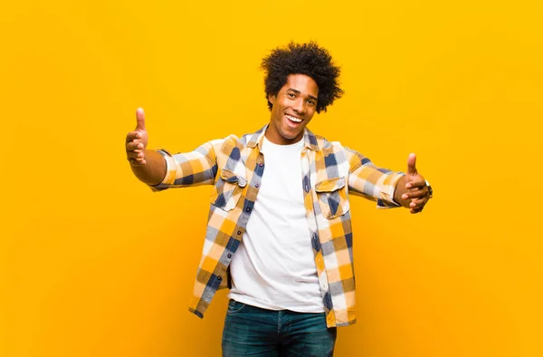 young black man smiling cheerfully giving a warm, friendly, loving welcome hug, feeling happy and adorable against orange wall