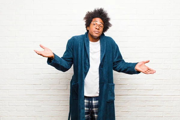 young black man wearing pajamas with gown feeling puzzled and confused, unsure about the correct answer or decision, trying to make a choice against brick wall