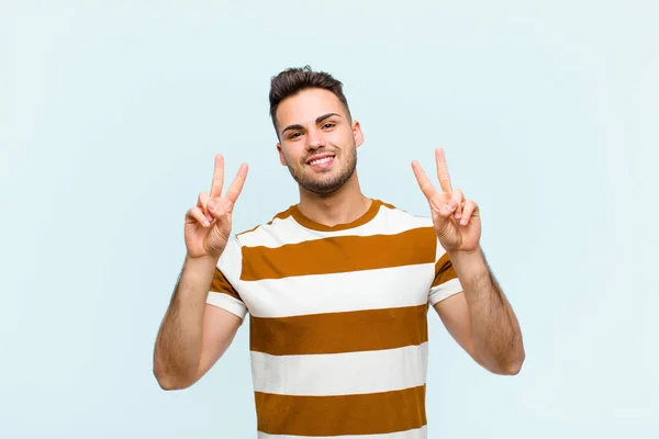若いヒスパニック系の男は笑顔で幸せそして友好的で満足している青い壁に対して両手で勝利や平和をジェスチャー — ストック写真