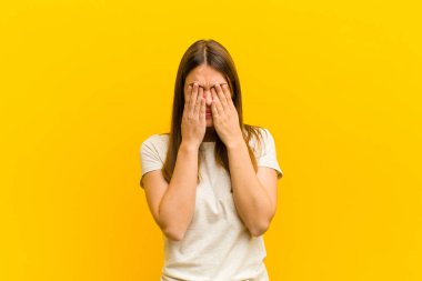 young pretty woman feeling sad, frustrated, nervous and depressed, covering face with both hands, crying against orange background