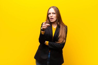 young pretty businesswoman pointing at camera with an angry aggressive expression looking like a furious, crazy boss against orange background