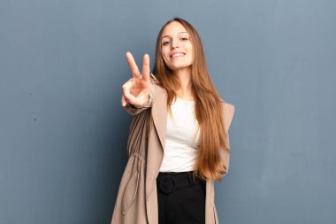 young pretty woman smiling and looking happy, carefree and positive, gesturing victory or peace with one hand against gray background