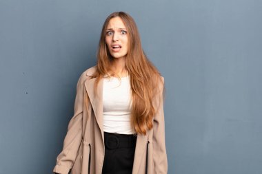 young pretty woman feeling terrified and shocked, with mouth wide open in surprise against gray background