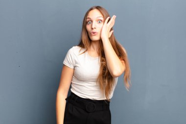 young pretty woman feeling shocked and astonished holding face to hand in disbelief with mouth wide open against gray background