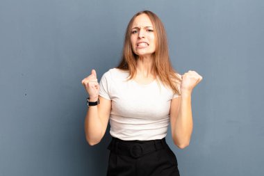 Genç ve güzel bir kadın sinirli bir ifadeyle ya da gri arka planda başarıyı kutlayan yumruklarla agresif bir şekilde bağırıyor.