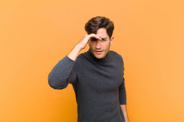 Genç yakışıklı adam şaşkın ve şaşırmış görünüyor, el üstünde uzak bakıyor, turuncu arka planı izliyor ya da arıyor