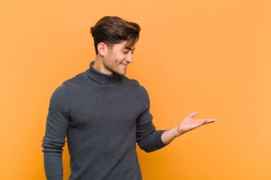 Genç, yakışıklı bir adam neşeyle gülümsüyor, mutlu hissediyor ve fotokopi alanında elini turuncu arka planda tutuyor.