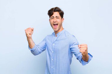 Genç yakışıklı adam muzaffer bir şekilde bağırıyor, heyecanlı, mutlu ve şaşırmış görünüyor, mavi arka planda kutlama yapıyor.