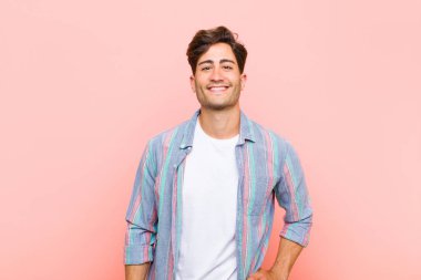 Genç yakışıklı adam mutlu bir şekilde gülümsüyor, eli kalçasında ve kendinden emin, pozitif, gururlu ve pembe arka plana karşı arkadaş canlısı.