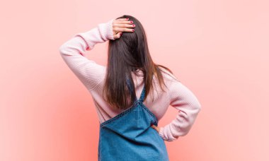 Genç güzel bir kadın düşünüyor ya da şüphe ediyor, kafasını kaşıyor, şaşkın ve şaşkın hissediyor, pembe arka planda ya da arka planda.