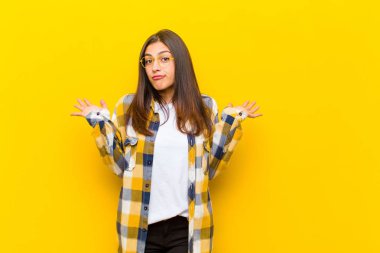 Genç, güzel bir kadın şaşkın ve şaşkın, şüpheci, farklı seçenekleri tartıyor ya da turuncu duvara karşı komik bir ifadeyle seçiyor.