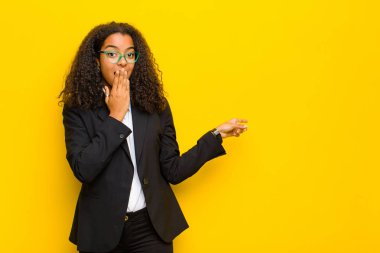 Siyah iş kadını kendini mutlu, şaşkın ve şaşırmış hissediyor. Ağzı elle kaplıyor ve turuncu duvara karşı yanal fotokopi alanını işaret ediyor.