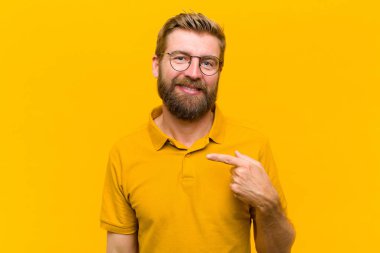 Genç sarışın adam gururlu, kendinden emin ve mutlu görünüyor, gülümsüyor ve kendini işaret ediyor ya da turuncu duvara karşı bir numara işareti yapıyor.