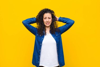 Genç, güzel bir kadın stresli, endişeli, endişeli ya da korkmuş, elleri başının üstünde, turuncu duvara karşı panikliyor.