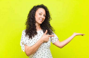 Genç, güzel bir kadın gülümsüyor, mutlu, kaygısız ve tatmin olmuş hissediyor, fotokopi alanındaki konsepte veya fikre işaret ediyor, kroma tuşlu duvara karşı.