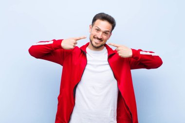 Genç yakışıklı sporcu ya da monitör kendi geniş gülüşünü işaret ediyor, pozitif, rahat, düz duvara karşı tatmin olmuş tavır.