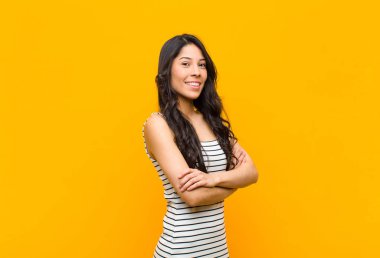 genç güzel latin kadın çapraz kolları ve mutlu, kendinden emin, memnun ifade, turuncu duvara karşı yanal görünümü ile kameraya gülümseyen