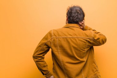 Genç yakışıklı adam düşünüyor ya da şüphe ediyor, kafasını kaşıyor, şaşkın ve kafası karışık hissediyor, turuncu duvarın arkasında ya da arkasında.