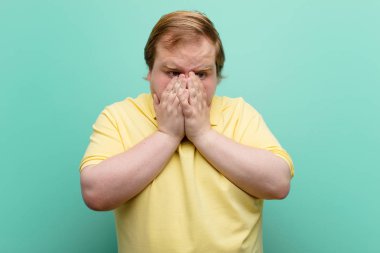 Genç, iri bir adam üzgün, hayal kırıklığına uğramış, gergin ve depresif, yüzünü iki eliyle kapatmış, mavi duvara karşı ağlıyor.