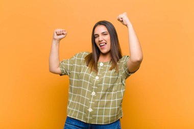 Genç, güzel bir kadın muzaffer bir şekilde bağırıyor, heyecanlı, mutlu ve şaşırmış görünüyor, turuncu duvarın karşısında izole bir şekilde kutluyor.