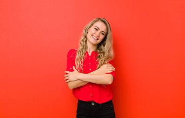 Genç sarışın kadın kollarını kavuşturup mutlu bir şekilde gülüyor, kırmızı duvara karşı rahat, pozitif ve tatmin olmuş bir pozla.