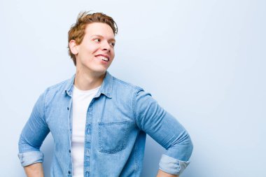 Genç kızıl saçlı adam mutlu, neşeli ve kendinden emin görünüyor. Gururla gülümsüyor ve iki elini de mavi duvara yaslamış kalçalarını kolluyor.