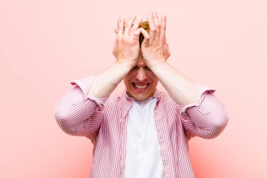 Genç kızıl saçlı adam stresli ve endişeli, depresif ve hayal kırıklığına uğramış hissediyor, iki elini de pembe duvara doğru kaldırıyor.