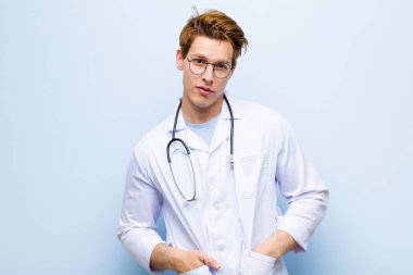 Genç kızıl saçlı doktor gururlu, kendinden emin, havalı, küstah ve küstah görünüyor, gülümsüyor, mavi duvara karşı kendini başarılı hissediyor.