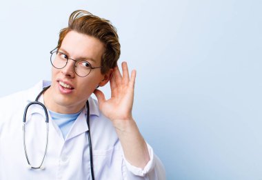 Genç kızıl saçlı doktor ciddi ve meraklı görünüyor, dinliyor, gizli bir konuşma ya da dedikodu duymaya çalışıyor, mavi duvara kulak misafiri oluyor.