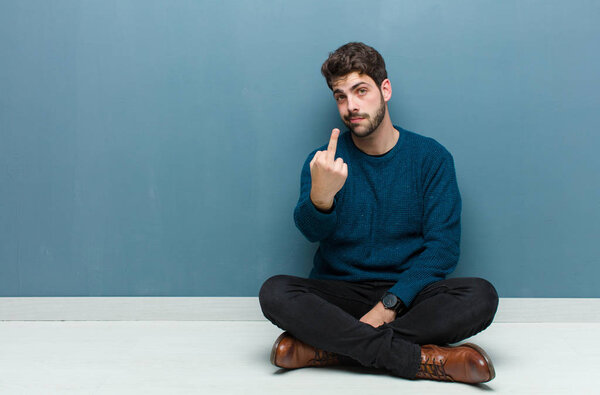 young handsome man sitting on floor feeling angry, annoyed, rebellious and aggressive, flipping the middle finger, fighting back