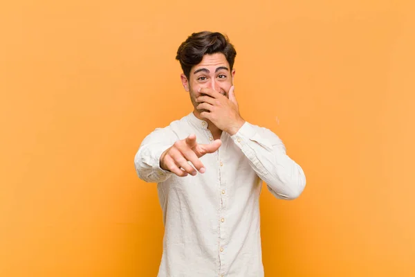 young handsome man laughing at you, pointing to camera and making fun of or mocking you against orange background