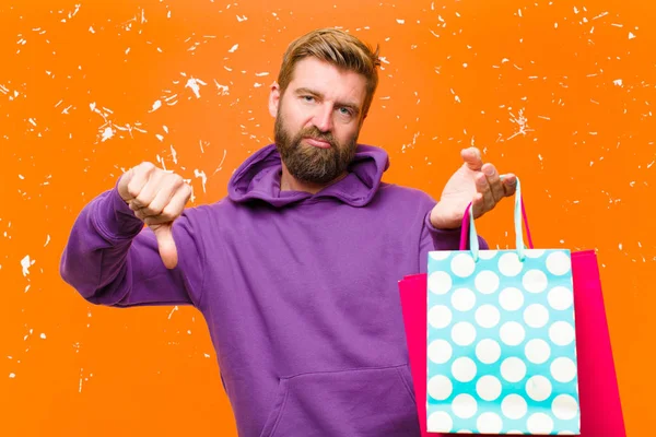 若いですブロンド男でショッピングバッグを身に着けています損傷したオレンジの壁に対して紫色のパーカー — ストック写真
