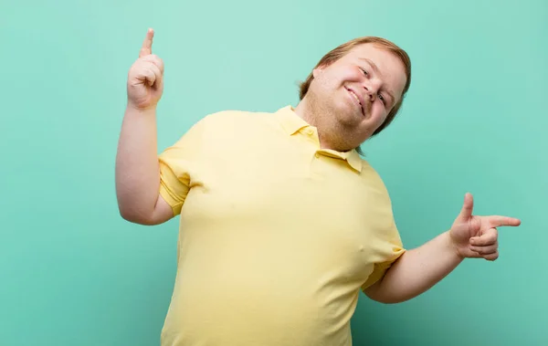 young big size man smiling, feeling carefree, relaxed and happy, dancing and listening to music, having fun at a party against blue wall