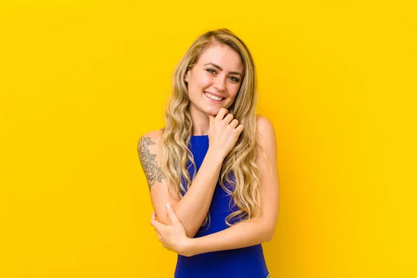 若いブロンドの女性は笑顔で人生を楽しんで幸せを感じ友好的で満足し屈託のない黄色の壁に対する顎の手で — ストック写真