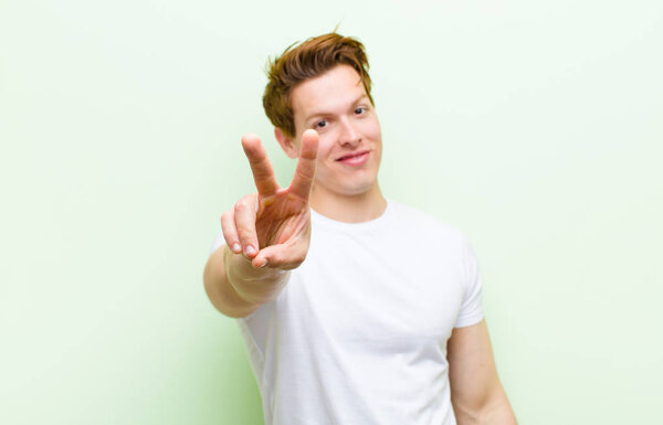 young red head handsome man smiling and looking happy, carefree and positive, gesturing victory or peace with one hand