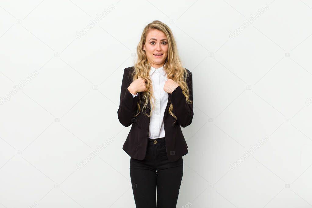 young blonde woman feeling happy, surprised and proud, pointing to self with an excited, amazed look against white wall