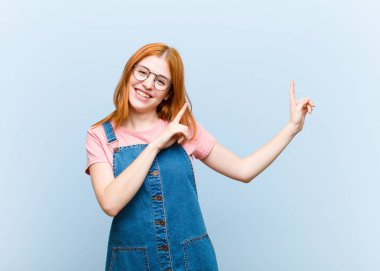 Genç kızıl saçlı güzel kadın mutlu bir şekilde gülümsüyor ve her iki elini de fotokopi uzayında nesne göstererek yukarı ve yana doğru gösteriyor.