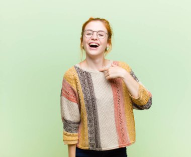 Genç, güzel kızıl saçlı kadın mutlu, gururlu ve şaşırmış görünüyor, neşeli bir şekilde kendini gösteriyor, kendinden emin ve yeşil duvara karşı yüce hissediyor.