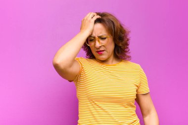 Orta yaşlı bir kadın mutsuz ve stresli görünüyor, intihar jesti elinde silah işareti yapıyor, kafasını mor duvara dayıyor.