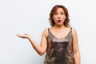 Orta yaşlı güzel bir kadın şaşırmış ve şok olmuş görünüyor, çenesi düşmüş beyaz duvara karşı eli açık bir nesne tutuyor