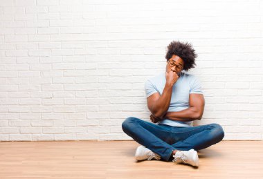 Genç siyah adam kendini ciddi, düşünceli ve endişeli hissediyor. Eli çenesine değmiş şekilde yan bakıyor. Evde yerde oturuyor.