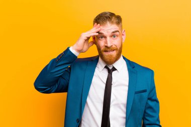 Genç kızıl saçlı işadamı mutlu, şaşkın ve şaşırmış görünüyor, gülümsüyor ve turuncu arka planda şaşırtıcı ve inanılmaz iyi haberleri fark ediyor.