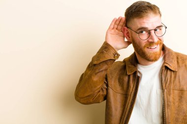 Genç kızıl saçlı adam gülümsüyor, tuhaf bir şekilde yan tarafa bakıyor, dedikoduları dinlemeye çalışıyor ya da bej arka plandaki bir sırrı duymaya çalışıyor.