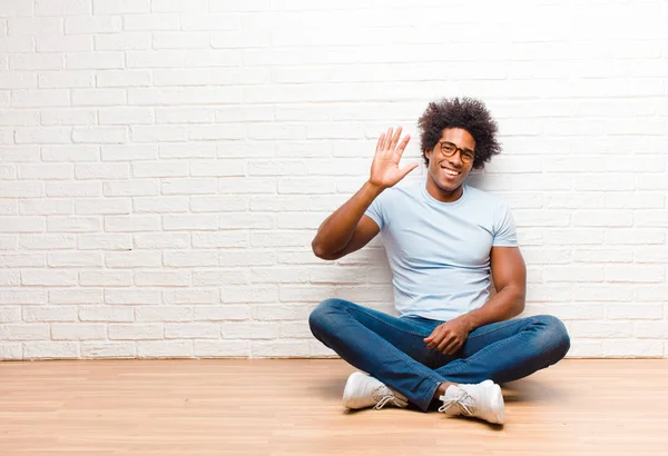 young black man smiling happily and cheerfully, waving hand, welcoming and greeting you, or saying goodbye sitting on the floor at home