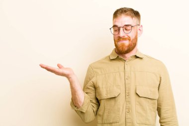 Genç kızıl saçlı adam mutlu ve gülümsüyor. Bej arka planda duran bir nesneye ya da konsepte bakıyor.