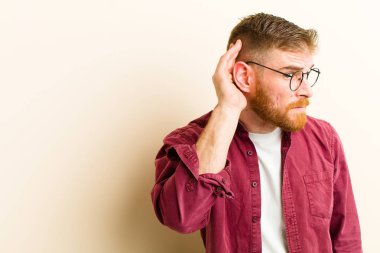 Genç kızıl saçlı adam ciddi ve meraklı görünüyor, dinliyor, gizli bir konuşma ya da dedikodu duymaya çalışıyor, bej arka plana kulak misafiri oluyor.
