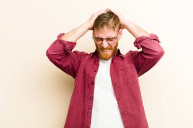 Genç kızıl saçlı adam stresli ve hayal kırıklığına uğramış hissediyor, el ele tutuşuyor, yorgun, mutsuz ve bej arka planda migreni var.