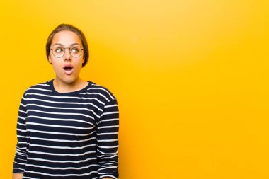 Genç, güzel bir kadın şok olmuş, mutlu, şaşkın ve şaşırmış hissediyor. Ağzı turuncu arka planda olan tarafa bakıyor.