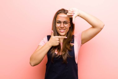 Genç bir kadın mutlu, arkadaş canlısı ve pozitif hissediyor, gülümsüyor ve pembe arka planda elleri olan bir portre veya fotoğraf çerçevesi yapıyor.