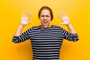 Genç, güzel bir kadın panik ya da öfke içinde bağırıyor. Şok olmuş, korkmuş ya da öfkeli. Elleri başının yanında turuncu arka planda.